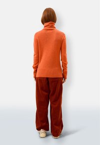 Oranges Rollkragenpullover aus strukturiertem Stoff, kombiniert mit rostfarbenen Cordhose und hellen Sneakers. Rückansicht.