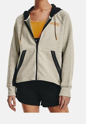 Personne portant un sweat à capuche beige zippé avec des poches en mesh noir et un short noir, debout devant un fond blanc uni.