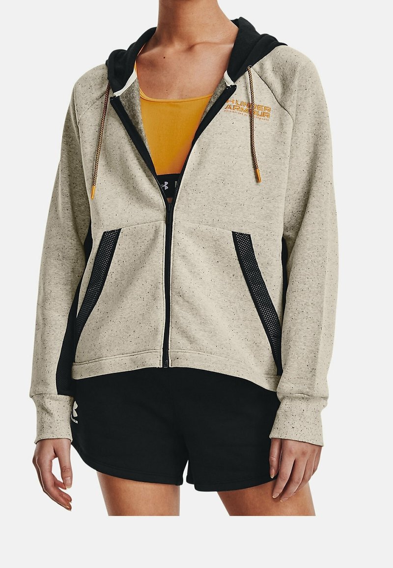 Personne portant un sweat à capuche beige zippé avec des poches en mesh noir et un short noir, debout devant un fond blanc uni.