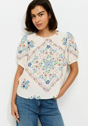 Vrouw die een crèmekleurige bloes met bloemenpatroon en pofmouwen draagt, gecombineerd met blauwe spijkerbroek, staand met één hand in de zak tegen een witte achtergrond.