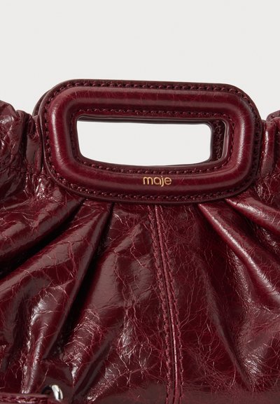 Gros plan d'un sac à main en cuir rouge foncé avec une poignée rectangulaire et un petit logo doré "maje" sous la poignée.