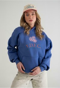 Blå hættetrøje med oversized pasform, frontlomme og blomsterdesign. Båret med en cremefarvet kasket med lyserøde detaljer og tekst.
