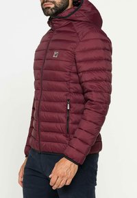 Giacca a vento maroon con cappuccio, caratterizzata da un design trapuntato, accento del logo argentato sul petto e tasche laterali con zip. Texture morbida.