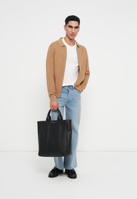 Fekete bőr bevásárlótáska függőleges formával, duplakezű fogantyúkkal és függőleges varrásokkal. A modellen bézs kardigán és világoskék farmernadrág található.