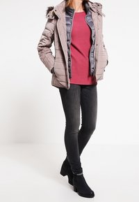 Gepolsterte graue Jacke mit einem kapuzenähnlichen Fellbesatz, darüber ein bordeauxrotes Oberteil, kombiniert mit dunklen Jeans und schwarzen Stiefeletten.