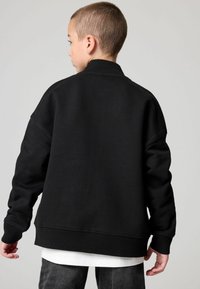 Felpa nera oversize con colletto e polsini a costine, caratterizzata da una texture liscia e spalle scese. Indossata sopra una camicia bianca.