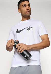 Fehér Nike póló fekete swoosh logóval, szürke sportnadrággal párosítva. Az illető egy fekete vizespalackot tart, ami csavaros kupakkal rendelkezik.