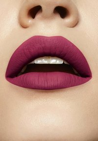 Lèvres bordeaux mates avec une finition texturée et un contour bien défini, dévoilant des dents blanches. Teint clair en arrière-plan.