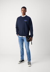 Mornarska majica s kapuljačom s logotipom, kombinirana s bijelom košuljom s ovratnikom, svijetloplavim trapericama, crnim tenisicama i crnim clutch torbicom.