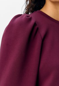 Bordeauxfarbene Bluse mit Puffärmeln, glatter Textur und Rundhalsausschnitt, die den Stofffall und die detaillierte Schulterraffung betont.