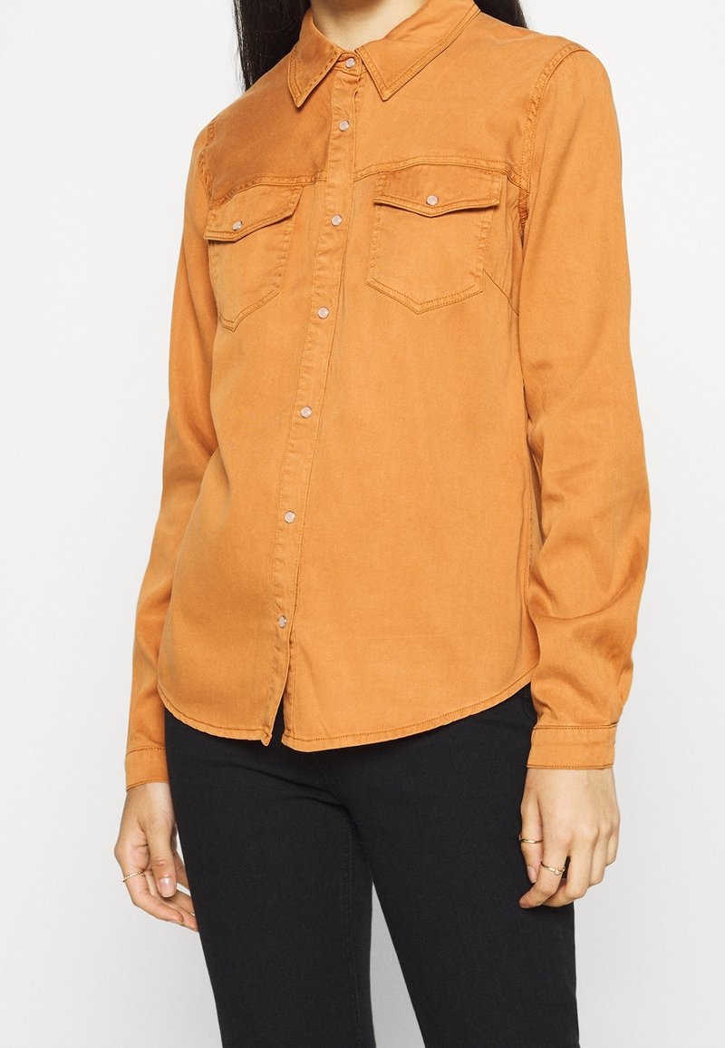 Femme portant une chemise orange à manches longues, boutonnée, avec deux poches poitrine, associée à un pantalon noir, debout devant un fond blanc.