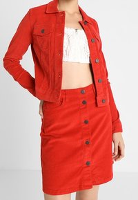 Giacca di corduroy rossa con tasche frontali e chiusura a bottoni, abbinata a una gonna camicia rossa coordinata. Tessuto liscio, design aderente.