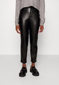 Osoba oblečená v černých kalhotách z umělé kůže, šedém pleteném svetru a černých chunky loaferech na jednobarevném světlejším pozadí.