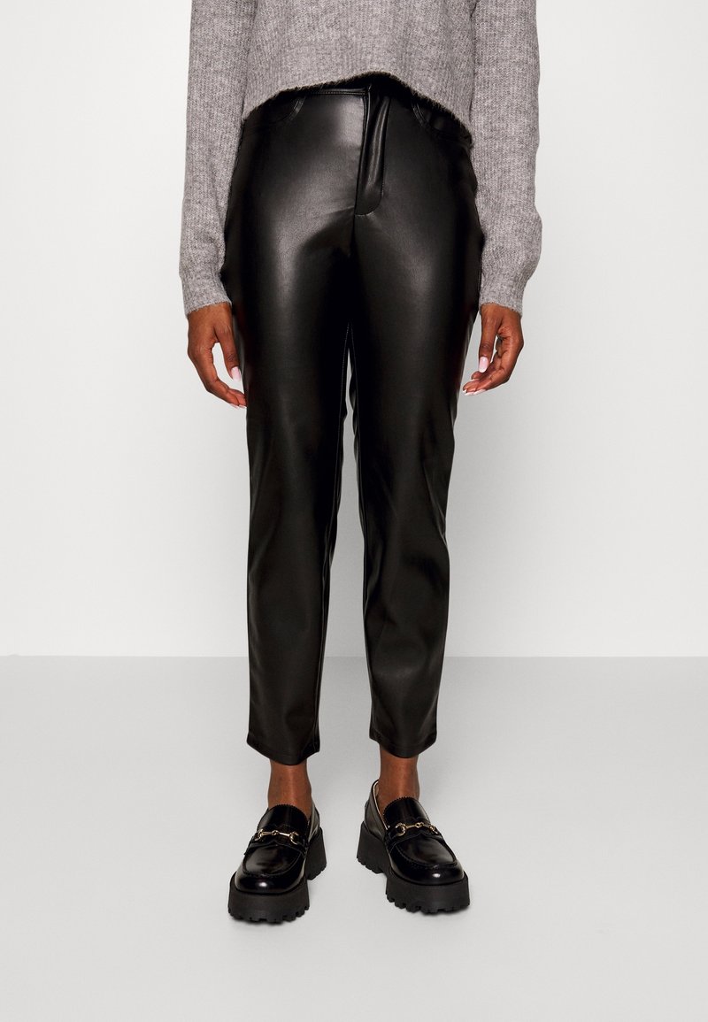 Osoba oblečená v černých kalhotách z umělé kůže, šedém pleteném svetru a černých chunky loaferech na jednobarevném světlejším pozadí.