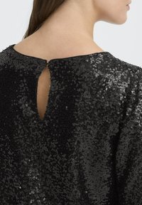 Musta paljettitoppi, jossa on selkäaukko, pyöristetty kaula-aukko ja teksturoitu pinta, joka hohtaa kimallusta.