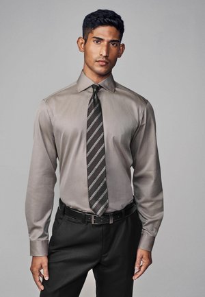 Camisa de vestir gris con mangas largas, cuello abotonado, combinada con una corbata oscura a rayas. Pantalones y cinturón negros completan el look formal.