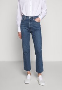 Jeans de mezclilla azul de talle alto con pierna recta y dobladillo deshilachado, combinados con una camisa blanca de botones y zapatillas blancas.