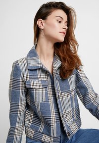 Giacca a quadri in blu, marrone e bianco. Realizzata in un tessuto testurizzato con colletto e tasche frontali. Chiusura con bottoni nella parte inferiore.