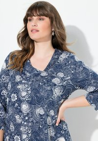 Frau mit welligem braunem Haar, die silberne Creolen und ein blaues, gemustertes Kleid mit weißen Blumen- und Wirbelmotiven trägt, posierend mit der Hand auf der Hüfte.