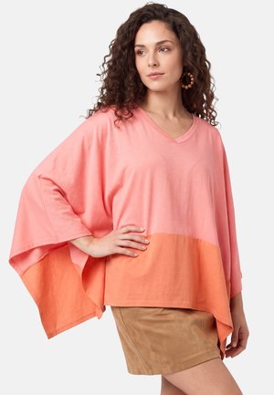Donna con capelli ricci che indossa una maglia larga rosa e arancione con maniche larghe e una gonna marrone chiaro, in piedi con la mano sul fianco.