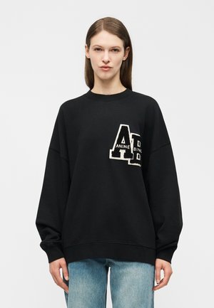 Γυναίκα με ίσια καστανά μαλλιά φοράει μαύρο φαρδύ φούτερ με μεγάλο μπαλώμα "AD" και ανοιχτόχρωμα μπλε τζιν, πάνω σε απλό φόντο.