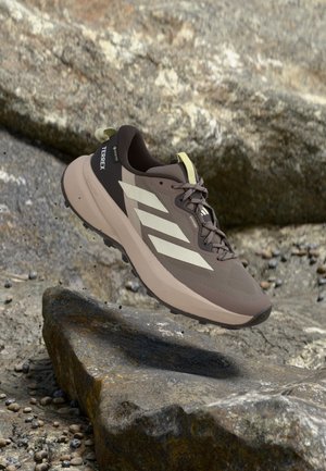 Zapatilla de trail running Adidas Terrex en tonos marrón y beige que flota sobre un terreno rocoso, con parte superior de malla y suela gruesa de goma.