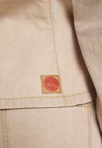 Tissu beige avec une texture subtile, présentant une patch logo rouge et des coutures jaunes le long de l'ourlet et des coutures.