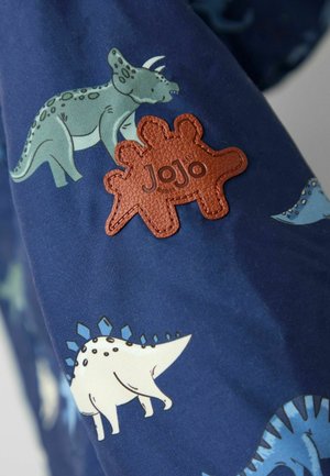 Modrá tkanina s barevnými vzory dinosauřích, s texturovanou hnědou koženou nášivkou ve tvaru tří dinosaurů, označenou "JoJo."