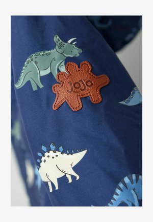 Plava tkanina s raznobojnim uzorcima dinosaura, uz teksturiranu smeđu kožnu oznaku u obliku tri dinosaura, označenu "JoJo."