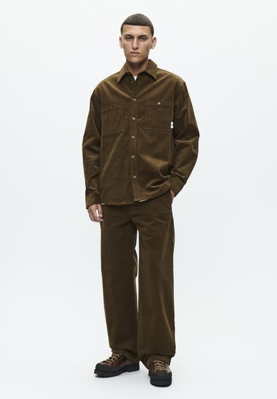 Brown corduroy shirt with two chest pockets paired with matching loose pants. The shoes are dark, with laces and chunky soles.