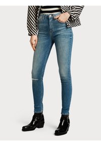 Kape slim vaaleansinisestä denimistä, jossa on kulunut pesu ja repeytyneet polviosat, yhdistettynä mustiin nilkkureihin.