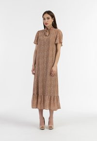 Maxikjole i brun stof med hvidt blomstermønster, korte ærmer, flagrende kant, høj hals med bindedetalje, og bæres med beige hæle.