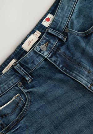 Niebieskie dżinsy o slim fit, z metalowym guzikiem i wieloma metkami z oznaczeniem rozmiaru oraz marki. Zawierają przednią kieszeń.