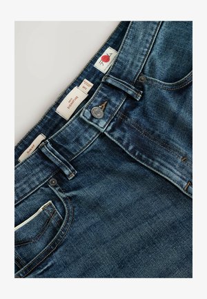 Niebieskie dżinsy o slim fit, z metalowym guzikiem i wieloma metkami z oznaczeniem rozmiaru oraz marki. Zawierają przednią kieszeń.