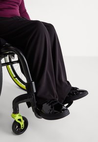 Μαυρο αμαξίδιο με ζωντανές πράσινες λεπτομέρειες; άτομο ντυμένο με μαύρο παντελόνι και γυαλιστερές μαύρες αθλητικές παπούτσια. Εστίαση στο κάτω μέρος του σώματος και στους τροχούς.