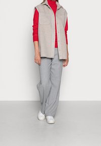 Grijze wollen vest met twee steekzakken aan de voorkant, gedragen over een rode coltrui, gecombineerd met lichtgrijze, wijd uitlopende broek en witte sneakers.