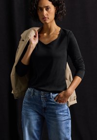 Schwarzes Langarmoberteil mit V-Ausschnitt, getragen mit blauen Jeans. Eine beige Jacke über einer Schulter drapiert.