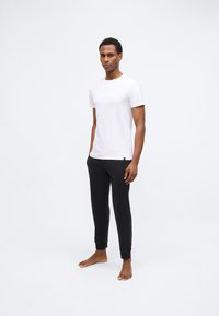 Férfi mezítláb, fehér, rövidujjú pólót és fekete sportnadrágot viselve, sima fehér háttér előtt.