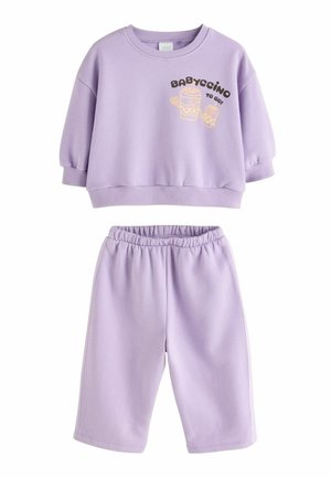 Småbarn med krøllet hår iført en lyse lilla sweatshirt med "Babyccino"-tryk, står udendørs ved en stenmur.
