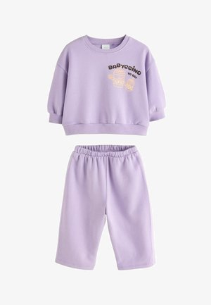 Småbarn med krøllet hår iført en lyse lilla sweatshirt med "Babyccino"-tryk, står udendørs ved en stenmur.