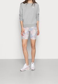 Woman standing wearing a gray Nike sweatshirt, pastel cloud-patterned bike shorts, and white and pink Nike sneakers on a gray floor.