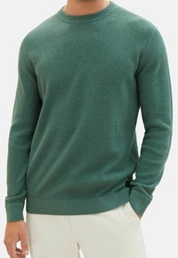 Grüner texturierter Pullover mit rundem Ausschnitt und langen Ärmeln, versehen mit gerippten Bündchen an Ärmeln und Saum. Dezentes Markenetikett an der Seite.