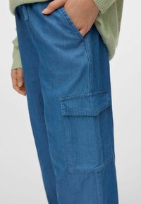 Pantalones cargo de mezclilla azul con bolsillos laterales y un corte relajado, que presentan costuras visibles y una textura suave. Combinados con un suéter verde claro.