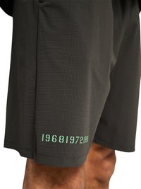 Zwarte textuurshorts gedragen door een persoon, met lichtgroene opgedrukte cijfers “1968 1972 1981” nabij de zoom op het linkerbeen.