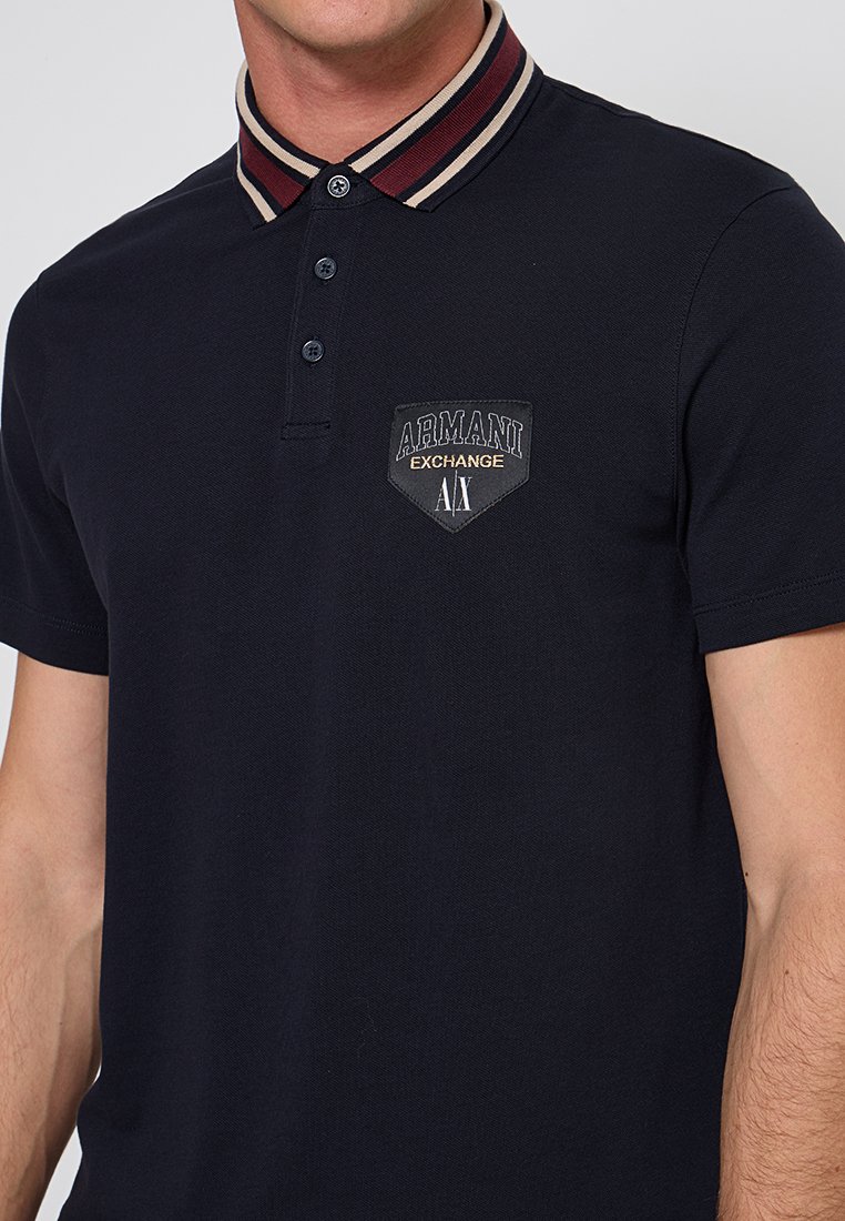 Marineblauw poloshirt met een gestreepte kraag in bordeauxrood en beige. Beschikt over een merklabel op de borst en een drieknops sluiting.