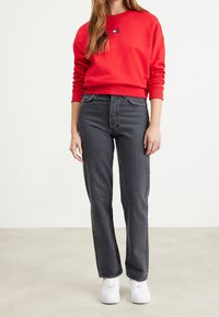 Rode sweatshirt met geribbelde mouwen, gecombineerd met hoogopgerichte grijze jeans met rechte pijpen. Witte sneakers maken de outfit af.