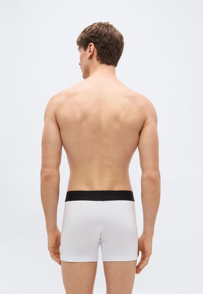 Άνδρας φαίνεται από πίσω φορώντας λευκά αντρικά εσώρουχα τύπου boxer με μαύρη ζώνη στη μέση, στέκεται μπροστά σε ένα απλό ανοιχτό γκρι φόντο.