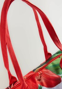 Rode tote bag met een levendig bloemenpatroon, voorzien van sterke geweven handgrepen en een ritsluiting. Het materiaal lijkt duurzaam en gestructureerd te zijn.