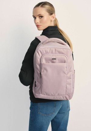 Liliowy plecak z wyściełanymi paskami i teksturowaną rączką, wyposażony w przednią kieszeń i boczne zamki, na tle czarnej puchowej kurtki.