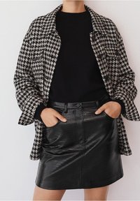 Mustavalkoinen houndstooth-kuvioinen takki mustan villatakin ja mustan tekonahkaisen mini-hameen päällä, jossa on etutaskut ja napitus.